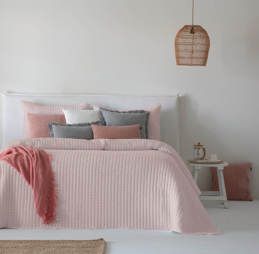 Colcha bouti lisa modelo Azara de color rosa nude sobre cama blanca con cojines decorativos a juego.